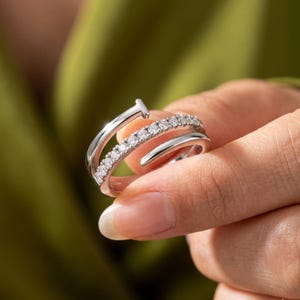 May include: A silver ring with a unique nail design. The ring features a band of small, sparkling stones and a curved, open design. The nail head is at the top of the ring. The ring is held in a hand against a blurred green background.
