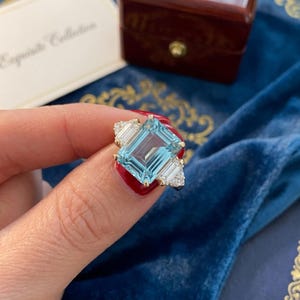 Peut inclure: Une bague en or ornée d'une grande pierre précieuse bleu clair taille émeraude en son centre. La pierre centrale est flanquée de deux pierres claires taille baguette de chaque côté, avec des pierres claires rondes plus petites sur l'anneau.