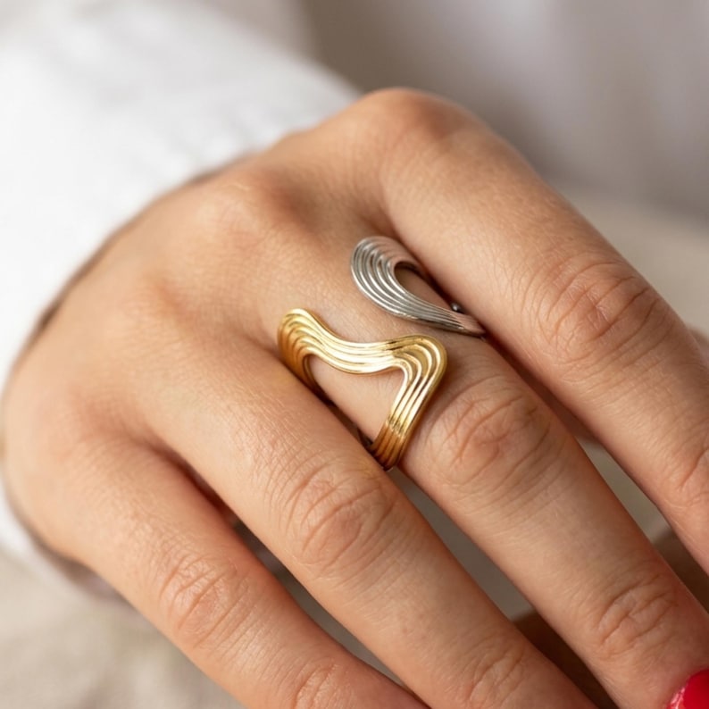 May include: A two-tone open ring featuring a gold-colored section and a silver-colored section. Both parts of the ring have a wavy, rippled texture with multiple parallel lines running along the length of the curved, open-ended design.