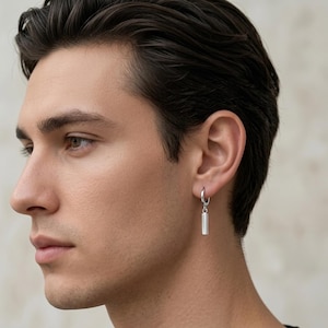 May include: A silver hoop earring with a rectangular bar pendant. The earring is worn on the right ear. The image is a close-up shot of the earring and the person's face.