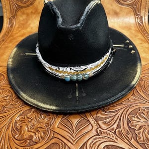 May include: A black fedora hat featuring a wide brim and a pinched crown. The hat is adorned with a decorative band consisting of white fabric with black patterns, a gold wavy trim, and three small, round, metallic blue-green beads at the front.