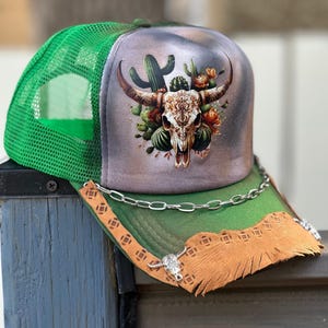May include: A green mesh trucker hat. The front panel is grey and green, featuring a bull skull design with cacti and flowers. The brim is green and brown with a chain and silver bull head accents.