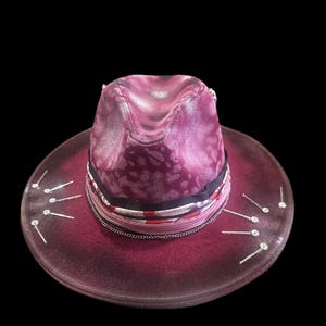 May include: A burgundy fedora hat with a textured crown and wide brim. The hat features a patterned band with a silver chain and decorative silver pins around the brim. The hat is set against a black background.