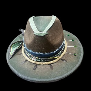 Peut inclure: Un chapeau fedora en feutre marron avec un bord vert foncé orné de paillettes noires. La calotte est décorée d'un ruban vert clair, d'une bande en tissu bleu effiloché, d'un ruban à motifs et d'une corde de jute. Des épingles vertes ornent le bord.