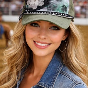 May include: An olive green trucker cap featuring a mesh back and a structured front panel. The front displays a white and green abstract swirl pattern, a black and white braided trim along the brim, and a large black, grey, and white crochet flower.