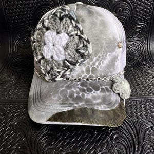 May include: A grey and white trucker hat with a crocheted flower detail. The hat features a silver chain and a decorative brim. The hat has a unique tie-dye pattern and a mesh back.