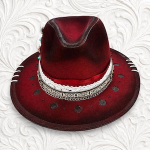 May include: A deep red fedora hat featuring a wide brim with white stitched accents and dark diamond-shaped cutouts. The crown is adorned with a white lace band, a patterned ribbon, a thin silver chain, and small turquoise and red bead details.