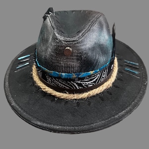 May include: A black fedora hat featuring a distressed finish, adorned with a tan jute rope band, a black and white paisley patterned fabric strip, and a vibrant blue fabric accent. A small metallic stud is centred on the front of the crown.