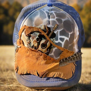 May include: A blue trucker hat with a mesh back and a decorative front panel. The panel features a guitar, a bull skull, and a brown leather fringe. A feather is attached to the side of the hat.