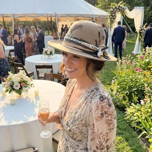 May include: A beige felt fedora hat featuring a dark botanical print, adorned with a layered ribbon band in shades of green, white, and black. The hat is paired with a long-sleeved, floor-length dress featuring a soft floral print pattern.