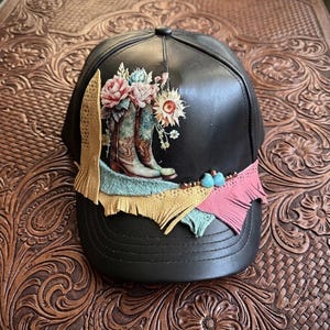 Può includere: Cappellino da baseball nero con un design floreale e stivali da cowboy. Il cappellino presenta una colorata disposizione di accenti rosa, blu e gialli, con una frangia in pelle sulla tesa. Il cappellino è appoggiato su una superficie marrone strutturata.