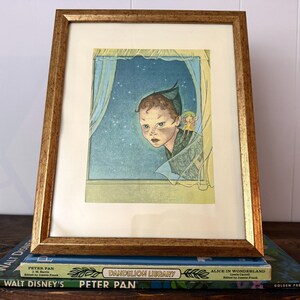 May include: A framed vintage illustration of Peter Pan looking out of a window at night, with a fairy. The artwork is in a gold-coloured frame and rests on a stack of books, including "Peter Pan" and "Alice in Wonderland".