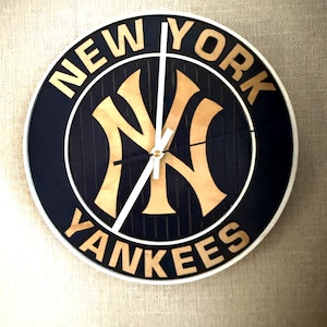 May include: A round wall clock featuring the New York Yankees logo in light wood tones against a dark blue background with vertical stripes. The text NEW YORK YANKEES is displayed in light wood lettering. It has white hour and minute hands.