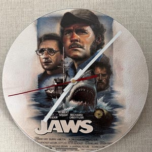 May include: A circular wall clock featuring the Jaws movie poster artwork, displaying the film title, a large shark emerging from the water, and portraits of the main characters. The clock has white hour and minute hands and a thin red second hand.