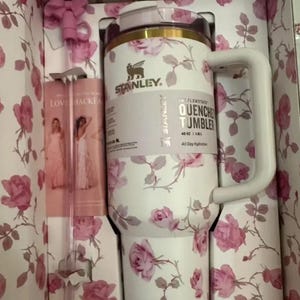 Puede incluir: Un vaso Stanley Quencher blanco con un estampado floral de rosas, presentado en una caja de regalo a juego. El vaso tiene un asa y una tapa. La caja tambi&eacute;n contiene una pajita rosa. El vaso es de 1,18L.