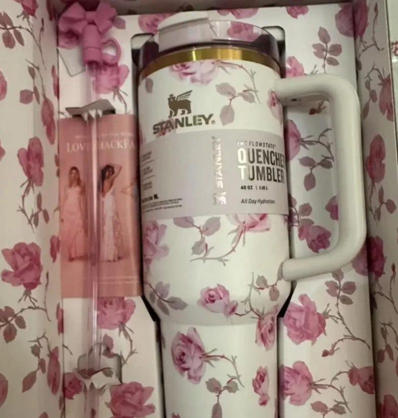 Puede incluir: Un vaso Stanley Quencher blanco con un estampado floral de rosas, presentado en una caja de regalo a juego. El vaso tiene un asa y una tapa. La caja tambi&eacute;n contiene una pajita rosa. El vaso es de 1,18L.