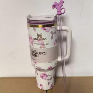 Puede incluir: Un vaso Stanley Quencher H2.0 blanco con un estampado floral de rosas y una pajita lila. El vaso tiene un asa y una tapa transparente. Los textos "Stanley" y "LOVESHACKFANCY" son visibles.