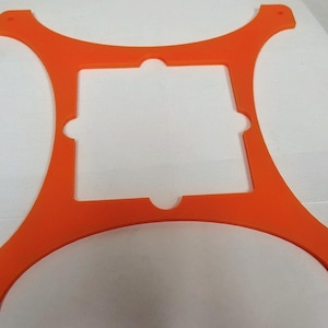 May include: Bright orange, four-armed bracket with a square center cutout. Each arm curves outward and has a small hole at the end. The bracket is made of a solid, opaque material.