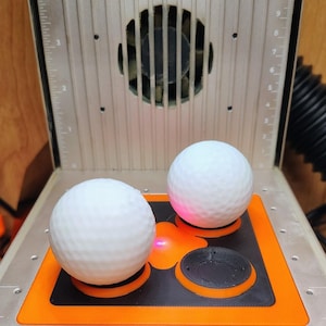 May include: Two white golf balls are positioned on an orange and black platform inside a laser engraving machine. The machine has a silver metal casing with a fan and measurement markings.