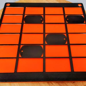 May include: A square black grid frame featuring a bright orange backing with sixteen rectangular slots. Three black, rounded rectangular tags are placed within specific slots of the grid. The frame has circular holes at each of the four corners.