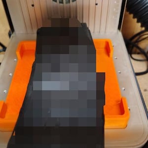May include: A 3D printer with an orange base and a black object inside. The printer is enclosed in a clear case, and the background includes a wooden surface and a black flexible tube.