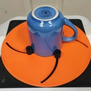 May include: A blue and purple ceramic mug with a handle, resting upside down on an orange circular stand. The stand has three adjustable black knobs and sits on a black square base. The mug has a speckled texture.