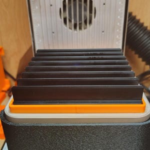May include: A close-up of a black and orange device with a series of black rectangular plates. The device has a textured black exterior and a light gray top with a fan and measurement markings. The plates are stacked inside the device.