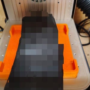 Könnte beinhalten: Ein 3D-Drucker mit orangefarbener Basis und einem schwarzen Objekt im Inneren. Der Drucker befindet sich in einem transparenten Gehäuse, der Hintergrund zeigt eine Holzoberfläche und einen schwarzen flexiblen Schlauch.