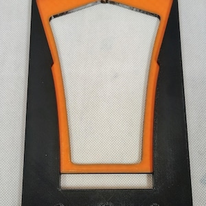 May include: A rectangular black plastic frame featuring a central orange trapezoidal insert. The black frame has a smooth finish with three circular holes aligned along the bottom edge, providing a structured mount for various mechanical components.