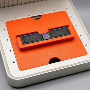 Puede incluir: Un dispositivo electrónico gris claro con una inserción rectangular de color naranja brillante. Centrado en la sección naranja hay un componente negro con el texto "DICE 478" y "300" impreso, acompañado por una pequeña luz indicadora de color violeta.
