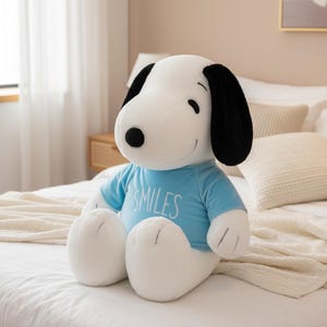 Puede incluir: Un peluche de Snoopy, principalmente blanco con orejas y nariz negras, que lleva una camiseta azul claro con la palabra "SMILES". El perro de peluche está sentado en una cama blanca, con una manta y almohadas color crema en el fondo.