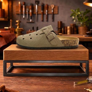 May include: A sage green clog with a buckle fastening and a cork footbed. The shoe has several small circular cut-outs and sits on a wooden display stand with a black metal frame. The background features tools in a workshop.