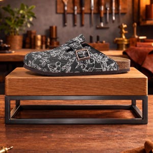 May include: A black and white patterned clog sandal with a buckle closure, displayed on a wooden stand with a metal frame. The sandal has a cork footbed and a dark brown sole. The background features tools and a wooden surface.