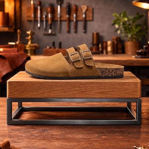 May include: A pair of brown suede clogs with three adjustable buckle straps, set on a wooden display stand with a metal frame. The clogs have a cork footbed and a black sole. The background features tools and a workshop setting.