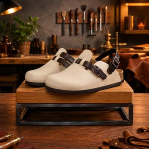 May include: A pair of cream-colored clogs with brown leather straps and buckles. The clogs have a cork sole and are displayed on a wooden stand with a black metal frame. The background features a workshop setting with tools.