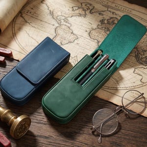 Op de afbeelding: Een gesloten blauwe lederen pennenetui en een open groene lederen pennenetui met drie metalen pennen. De groene etui heeft een klepsluiting en een speciaal binnenvak voor schrijfinstrumenten. Ronde metalen bril ligt in de buurt.