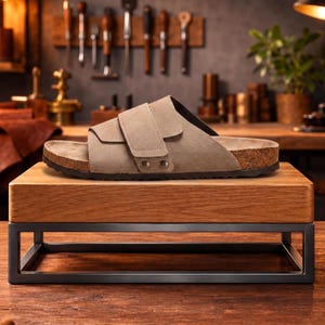 May include: A light brown suede sandal with two adjustable straps and a cork footbed. The sandal is displayed on a wooden block with a black metal frame, showcasing footwear in a studio setting.