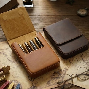 Puede incluir: Un estuche para bolígrafos de cuero color canela con la tapa abierta que revela cuatro ranuras individuales con bolígrafos metálicos, junto a un estuche de cuero marrón oscuro cerrado. El interior del estuche abierto tiene una textura suave.