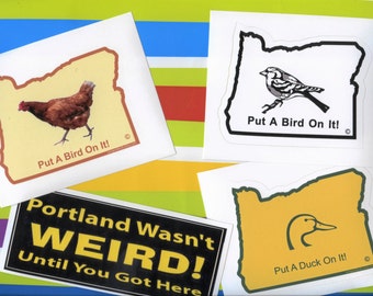 Op de afbeelding: Drie stickers met de omtrek van de staat Oregon met verschillende slogans en afbeeldingen. De eerste sticker toont een kip en de tekst "Put A Bird On It!". De tweede sticker toont een vogel en de tekst "Put A Bird On It!". De derde sticker toont een eend en de tekst "Put A Duck On It!".