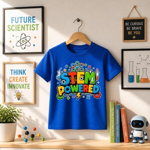 Könnte beinhalten: Ein blaues T-Shirt mit den Worten "STEM POWERED" in bunten Buchstaben und wissenschaftlichen Grafiken. Das Shirt hängt an einem Holzbügel. Gerahmte Kunst mit den Worten "FUTURE SCIENTIST" und "THINK CREATE INNOVATE" befindet sich im Hintergrund.
