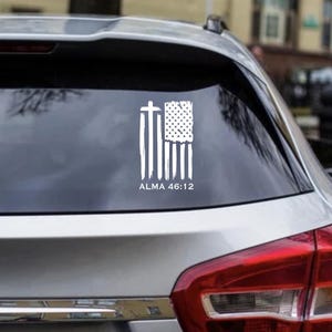 Puede incluir: Calcomanía de vinilo blanco con un diseño de bandera estadounidense desgastada con una cruz y el texto "ALMA 46:12." La calcomanía está aplicada en la ventana trasera de un coche plateado. El diseño combina símbolos religiosos y patrióticos.