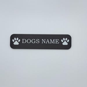 May include: A black rectangular sign with rounded corners, featuring the text "DOGS NAME" in white capital letters. White paw prints flank the text. The sign is designed for pet owners.