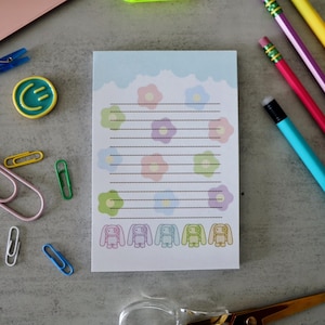 May include: A notepad with a light blue scalloped border, floral and bunny illustrations, and lined pages. Pencils, paperclips, and a laptop are scattered around the notepad on a gray surface.