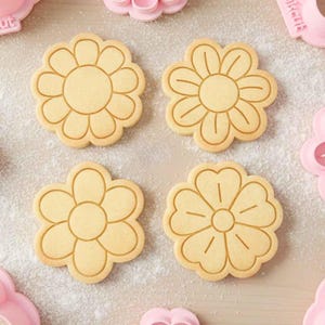 Pode incluir: Quatro biscoitos em forma de flor com desenhos detalhados de pétalas. Os biscoitos são castanhos claros com linhas gravadas, sugerindo uma textura assada. Cortadores de biscoitos rosa são visíveis no fundo, sugerindo um tema de cozimento.