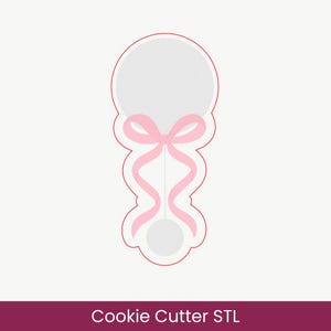 Pode incluir: Um cortador de biscoitos em forma de chocalho de bebé com um topo circular e uma base mais pequena ligados por uma linha vertical fina. O design apresenta um laço cor-de-rosa claro com duas fitas longas e fluidas atadas ao cabo.