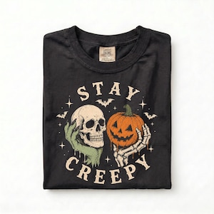 May include: A black t-shirt featuring a graphic print with the text STAY CREEPY. The design includes a beige human skull held by a green hand and an orange jack-o'-lantern held by a white skeletal hand, surrounded by small stars and bats.