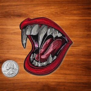 Puede incluir: Parche bordado que representa una boca abierta con afilados colmillos, una lengua roja y labios rojos. El parche tiene un fondo negro y está delineado en rojo. Una moneda de veinticinco centavos está en la esquina inferior izquierda.