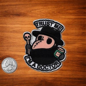 May include: Embroidered patch featuring a cartoon plague doctor with a black hat and mask, holding a staff and a small container. The patch has the text "TRUST ME I'M A DOCTOR" in white lettering with a black border.