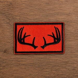 May include: A rectangular embroidered patch featuring a vibrant red background with a black border. Centered on the patch is a black graphic illustration of a pair of deer antlers, symmetrically arranged to face each other in the middle.