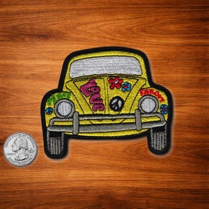 May include: Yellow embroidered patch of a classic car with the word "LOVE" in pink, a peace symbol, and flower accents. The car has a black outline and gray bumper. The patch is on a wooden surface.